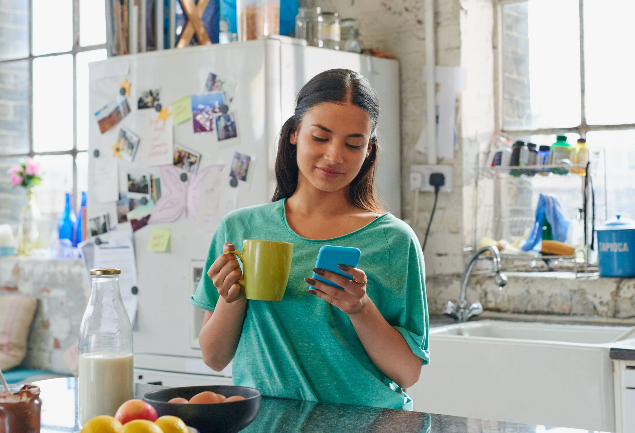 Woman drinking coffee in her kitchen while using her phone